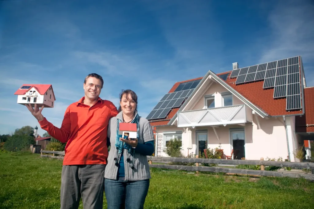 Glückliches Paar hält ein Modellhaus, im Hintergrund ihr Eigenheim mit großer Photovoltaikanlage